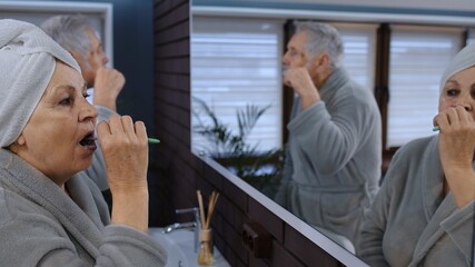 Cheerful old senior couple grandparents man and woman brushing teeth and looking into a mirror....