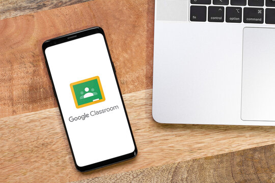 Smartphone displaying Google Classroom logo app, illustrative content of Google Classroom on a phone with top view of a wooden desk background