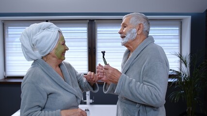 Old senior couple man and woman in bathrobe are dancing in bathroom having fun laughing enjoying leisure time and music. Elderly grandmother with facial green mask and grandfather with shaving foam
