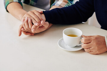 Close-up image of caring son holding hand of his mother who is drinking cup of hot tea