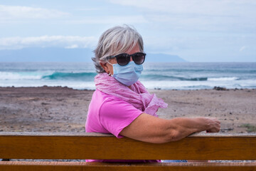 Coronavirus and new normal life. Senior woman looking into camera sitting on a bench wearing a surgical mask due to coronavirus while enjoying the day at the beach. Waves and horizon over water