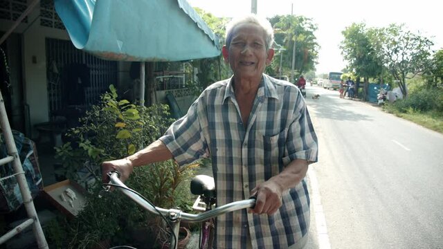 Slow motion of white-haired poor Asian elderly man , older than seventy stands smiling and is happily leading the bicycle on the roadside of his village