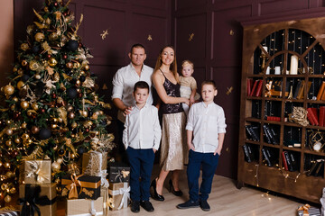 Fototapeta na wymiar A beautiful happy young family of five stands in full height near the Christmas tree and look into the camera. mother, father and three children