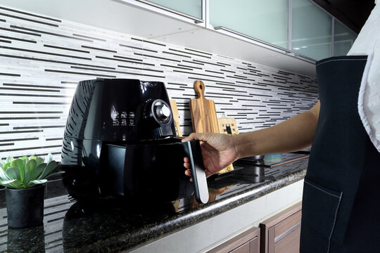 a right hand of lady woman is holding the tray of the air fryer or oil frying machine which is on the marble table of the modern interior design kitchen