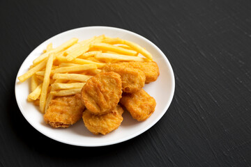 Tasty Fastfood: Chicken Nuggets and French Fries on a plate on a black surface, side view. Copy space.