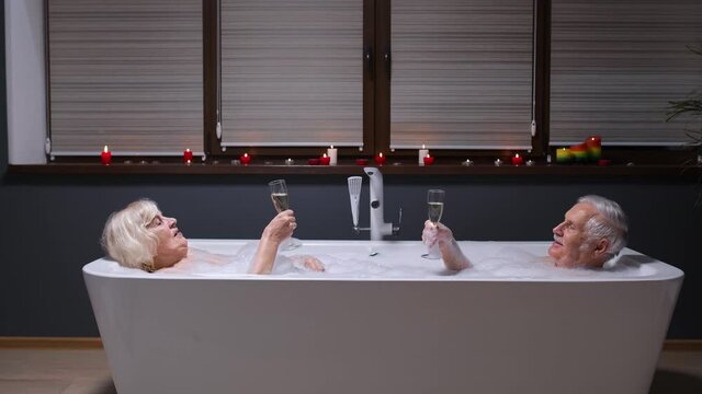 Sexy senior couple grandfather and grandmother is taking foamy bath, drinking champagne in luxury bathroom with candles. Elderly man, woman drinks wine. Spa procedures. Honeymoon. Life of retirees