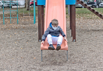 Playful kid with face mask having on a slide at the playground.