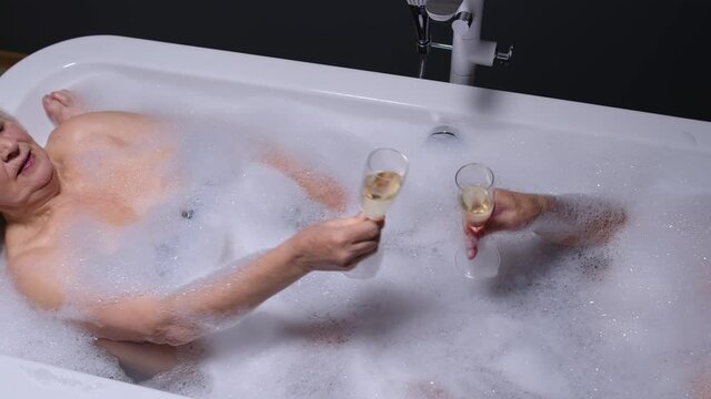 Sexy senior couple grandfather and grandmother is taking foamy bath, drinking champagne in luxury bathroom with candles. Elderly man, woman drinks white wine. Spa procedures in hotel. Honeymoon