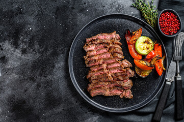 Sliced Grilled beef marbled rare steak. Chuck eye roll on a plate with a side dish of vegetables. Black background. Top view. Copy space