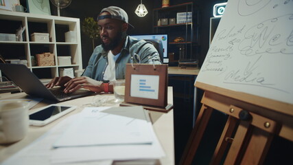 Fototapeta na wymiar Focused african american man reading news on laptop computer in dark coworking.