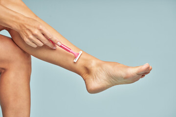 Fototapeta premium Depilation. Close up of lady shaving her leg with safety razor isolated on blue background