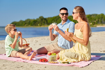 family, leisure and people concept - happy mother, father and little son having picnic on summer beach and eating sandwiches