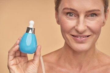 Beautiful mature happy woman holding the bottle of anti-aging serum and smiling at camera