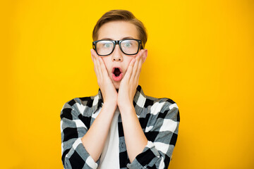 Portrait of teen boy shocked facial expression, holding hands near face and open mouth on yellow...