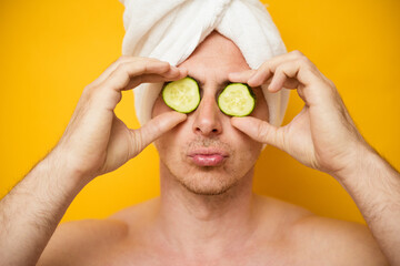 Headshot of funny guy, covers eyes with slices of cucumbers, has spa treatments, poses indoor. Men, beauty, cosmetology concept