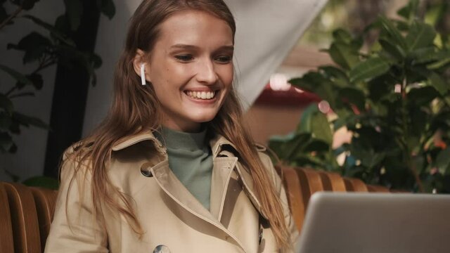 Beautiful happy student girl in wireless earphones waving hello having lecture by video call on laptop studying online in cafe outdoor. Greeting expression