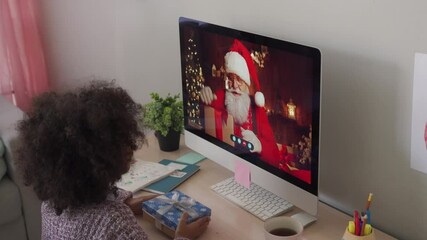 Happy african american kid girl talking to virtual santa claus saying New Year greetings to child on video conference online call chat using computer sitting at home table opening Chrtistmas present.