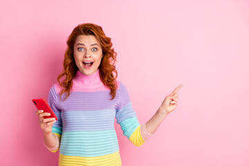 Photo portrait of excited woman holding phone in one hand pointing finger at blank space isolated on pastel pink colored background