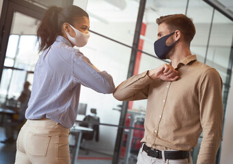 Two young diverse business colleagues wearing face protective masks bumping elbows, greeting each...