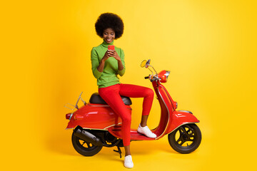 Fototapeta na wymiar Photo portrait of young brunette african american woman sitting on red scooter holding cellphone sending message wearing wearing green jumper isolated on vivid yellow colroed background