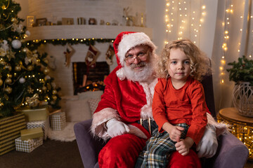 Fototapeta na wymiar Funny Santa Claus sitting on his rocker with little cute boy sitting on his knee. Christmas spirit, holidays and celebrations concept