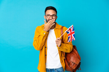 Young caucasian man holding an United Kingdom flag isolated on yellow background happy and smiling covering mouth with hand