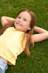 Dreaming... Portrait of joyful little girl in casual clothes lying on the green lawn, looking up and smiling while visiting park on a sunny summer day