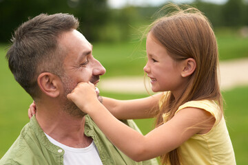 Close up of adorable little girl having fun with her loving daddy, touching his face while he is holding her and smiling, standing in the green park