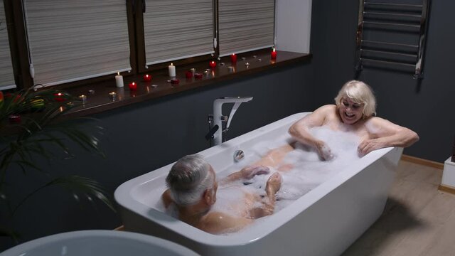 Sexy senior couple grandfather and grandmother is taking foamy bath, drinking champagne in luxury bathroom with candles. Elderly man, woman in love. Spa procedures in hotel. Honeymoon. Slow motion