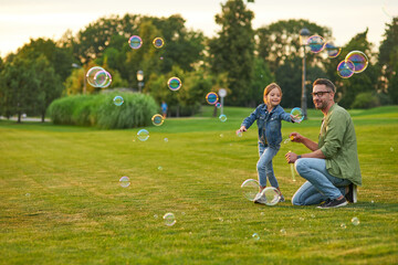 Full length shot of young father and his cute little girl having fun while blowing soap bubbles on a summer day, daughter and dad spending time together outdoors - Powered by Adobe