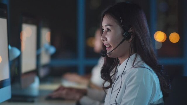 Millennial Asia young call center team or customer support service executive using computer and microphone headset working technical support in late night office. Telemarketing or sales job concept.