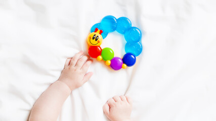 Baby hands playing with colorful rattle on white bed. Closeup. Toys of development for infant. Top view.