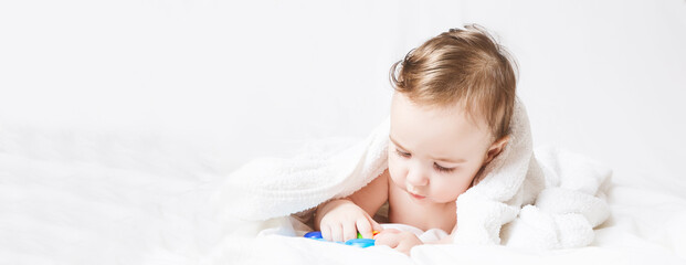 Obraz na płótnie Canvas Six months old baby wearing a towel after a bath. Baby care concept. Cute toddler playing with rattle on white bed. copy space. Banner