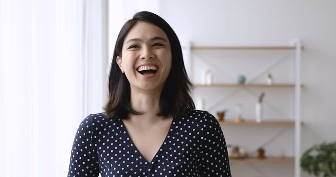 Happy young korean mixed race pretty woman laughing at funny joke, looking at camera, feeling excited of hearing good news, showing positive mood emotions, standing indoors in own apartment home.