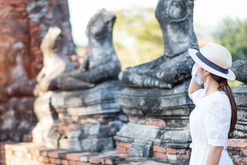 Tourist Woman in white dress wearing surgical face mask, protection COVID-19 pandemic during visiting in Wat Chaiwatthanaram temple in Ayutthaya. new normal, safety travel and Thailand travel concept