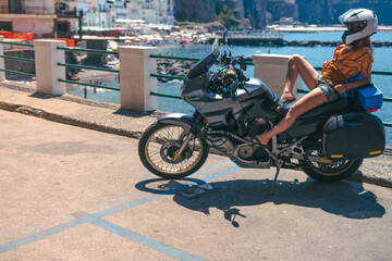 Young sexy girl posing. Motorcycle for tourism and travel. Wearing a helmet. In the background are views of the sea and hotels. Coast of Italy. Sunny day, vacation and trip.