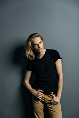 Young attractive guy with long blonde hair is sad leaning against a gray wall, looks to the camera