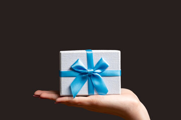 Holiday present. Festive surprise. Special occasion greeting. Female hand holding blue gift box on open palm isolated on dark brown copy space background.