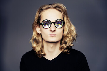 Young handsome guy with long blonde hair in a black T-shirt on a gray background with glasses...