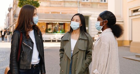 Fototapeta premium Three mixed-races young women, best friends meeting at city street and greeting with elbows during coronavirus pandemic. Multi ethnic females in medical masks talking cheerfully outdoor.