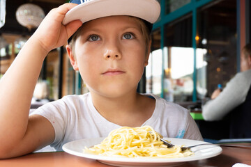 a boy in a cap 6 years old is sitting in a cafe and does not want to eat spaghetti, looking directly at the camera