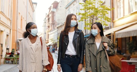 Three mixed-races young stylish women in medical masks talking and walking the street in city. Pretty cheerful multi ethnic females friends strolling outdoors and watching town. Tourists at pandemic.