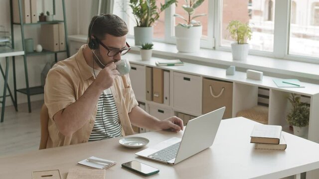 Medium high angle of brown-haired Caucasian man wearing headset sitting by desk at home office, drinking from cup, talking on business call. Worker using laptop, taking notes with pen