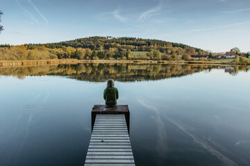 Fototapeta premium Calm morning relaxation by a pond. Sitting woman in the countryside. Intended female mysterious atmosphere. Spring sunny afternoon in the nature. Silence. Wooden pier on a pond. View of a fresh nature