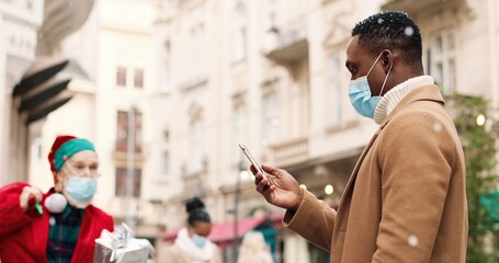 Cheerful old Caucasian Santa Claus with bag full of presents giving gift box to happy surprised African American male in mask who is texting on smartphone. Man meets Santa on street in x-mas city