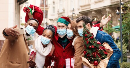 Portrait of happy African American and Caucasian men and women friends in masks with presents in city and talking while meeting old Santa Clause. Santa giving gifts to surprised female. X-mas concept