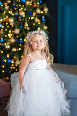 Cute little girl blonde with blue eyes, looking like an angel in a white dress, near the Christmas tree