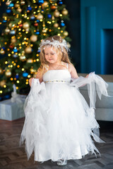 Cute little girl blonde with blue eyes, looking like an angel in a white dress, near the Christmas tree