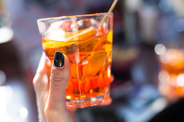 Woman holds a glass with orange spritz in sunny day. Hand with black nails. Serving drinks outdoors. Isolation concept. Traditional venetian italian cocktail. Celebration concept. Spirits take-away. 