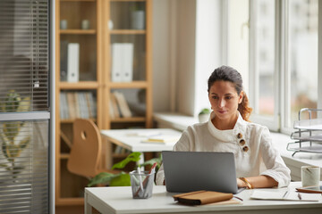 Wide angle portrait of modern businesswoman using laptop while sitting at desk by window in office, copy space